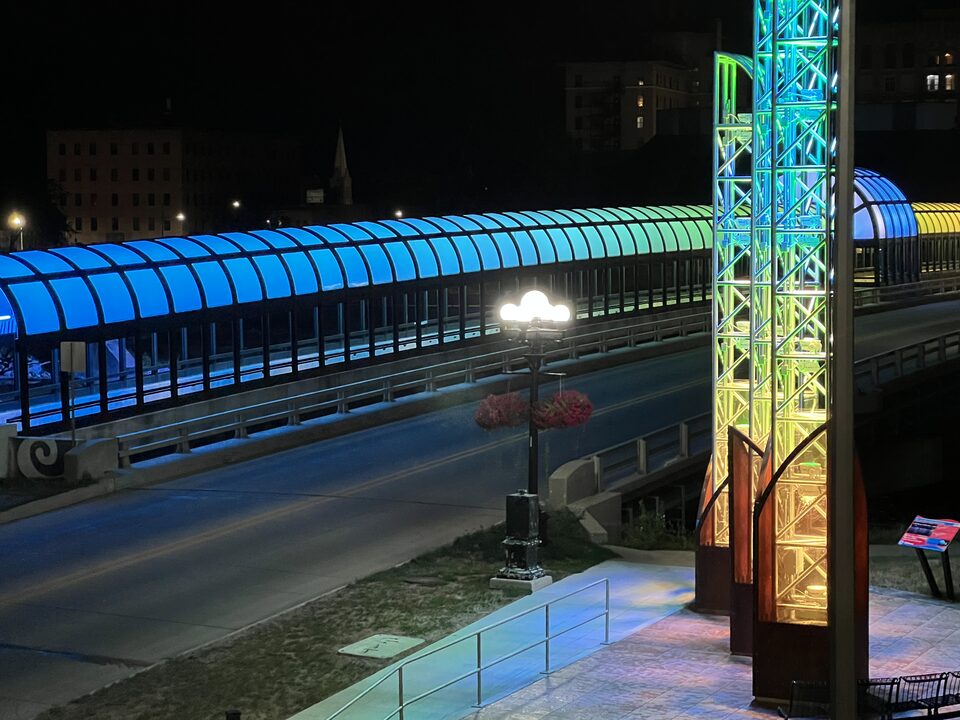 An enclosed pedestrian skywalk lit at night over the Cedar River in downtown Waterloo, Iowa
